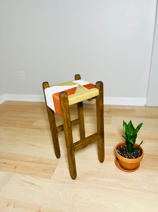 A wooden stool with a colorful woven seat sits on a wooden floor, accompanied by a potted plant in the background.