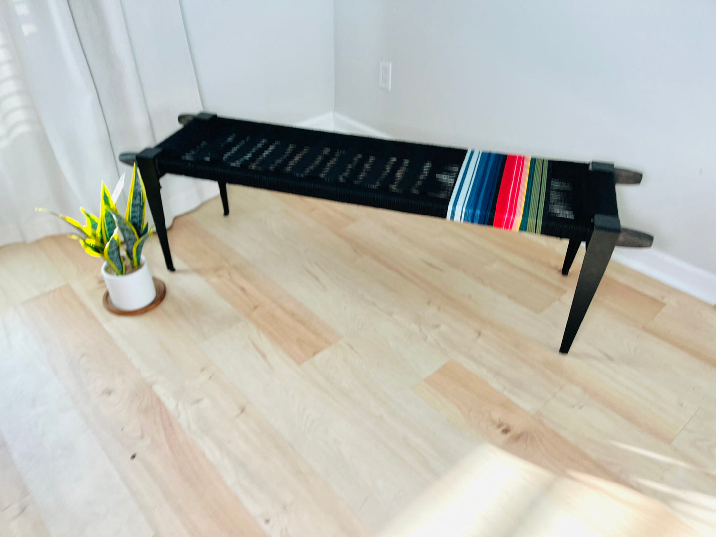 a black bench with colorful stripes on the armrest, placed on a wooden floor next to a potted plant.