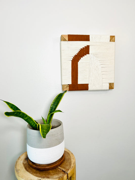 a potted plant with green leaves and a wooden stool with a woven basket, set against a white wall with a framed artwork hanging above.