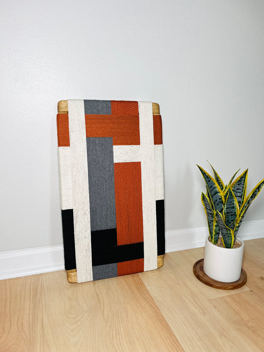 a wooden floor with a colorful woven piece of art, a potted plant, and a white vase on a wooden stand.