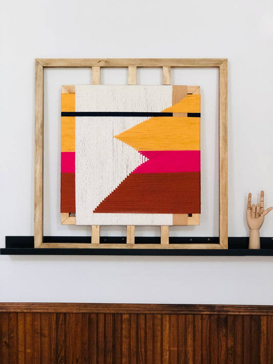 a wooden shelf with a framed abstract artwork hanging on it. The artwork is a colorful geometric design with a mix of pink, orange, and white colors. The wooden shelf has a black base and is positioned against a white wall.