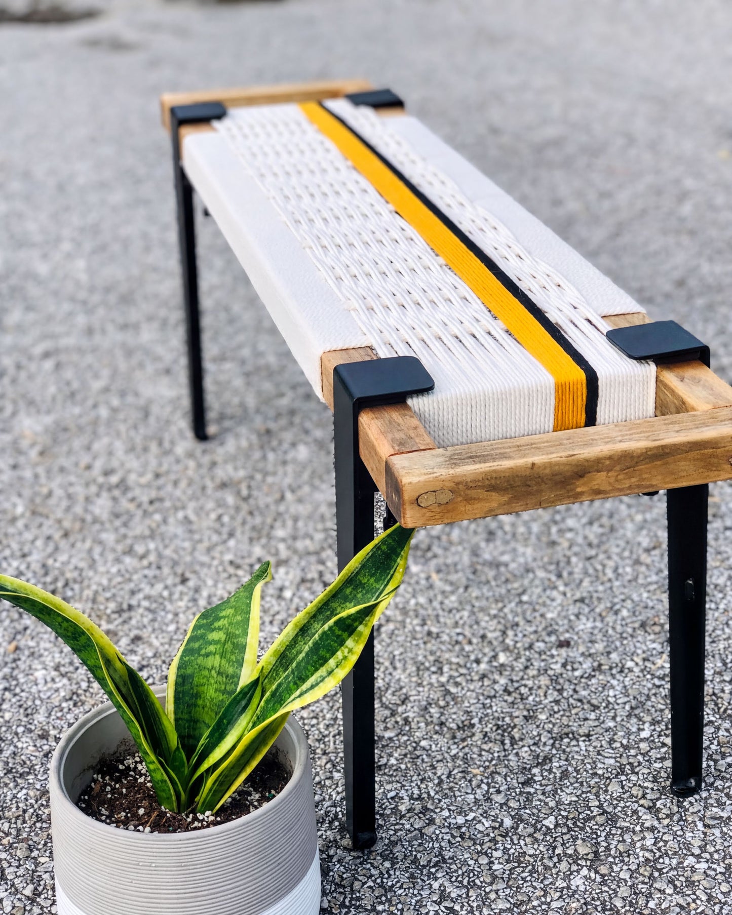 a wooden bench with a woven seat and metal legs, placed on a gravel surface next to a potted plant.
