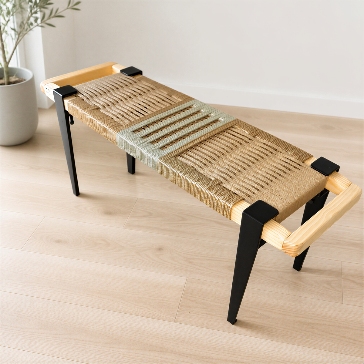 A woven bench with a wooden frame and black metal legs, placed on a wooden floor against a white wall.