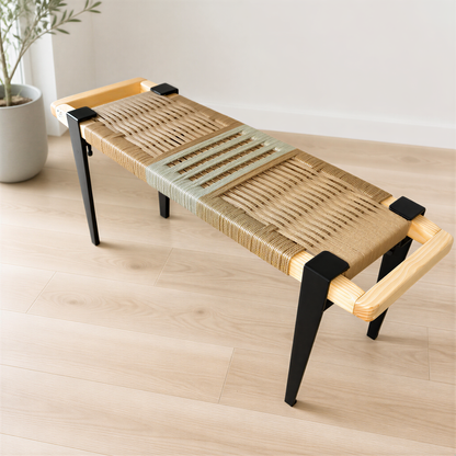 A woven bench with a wooden frame and black metal legs, placed on a wooden floor against a white wall.