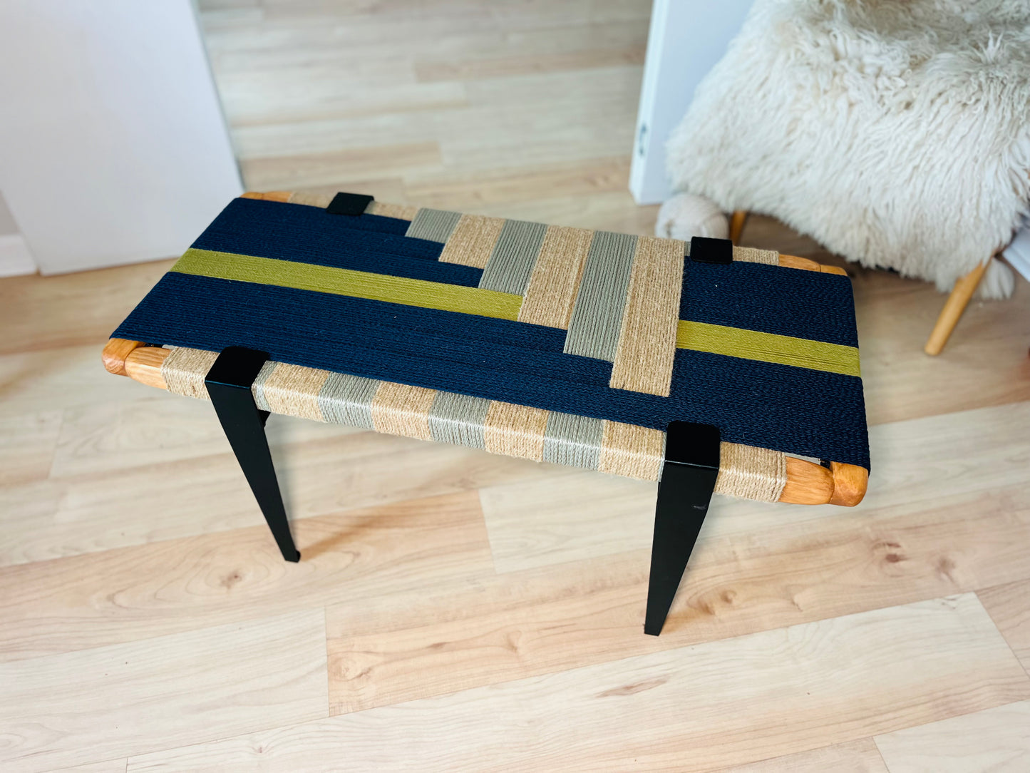 a modern, handcrafted coffee table made of woven materials in shades of blue, yellow, and gray. The table has a unique, geometric design with a striped pattern and a wooden base. The table is placed on a wooden floor, and there is a white fur rug visible in the background.