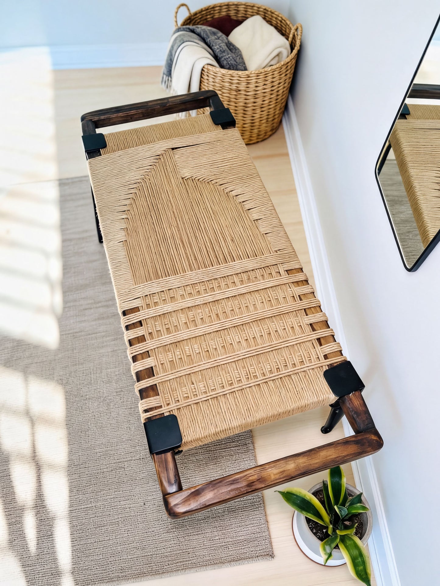 a woven bench with a wooden frame, placed on a rug in a room with a basket and a plant nearby.