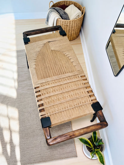 a woven bench with a wooden frame, placed on a rug in a room with a basket and a plant nearby.