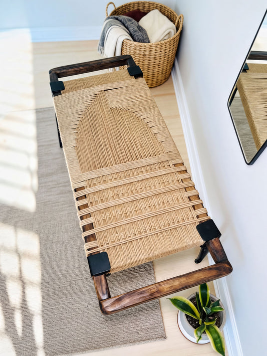 a woven bench with a wooden frame, placed on a rug in a room with a basket and a plant nearby.