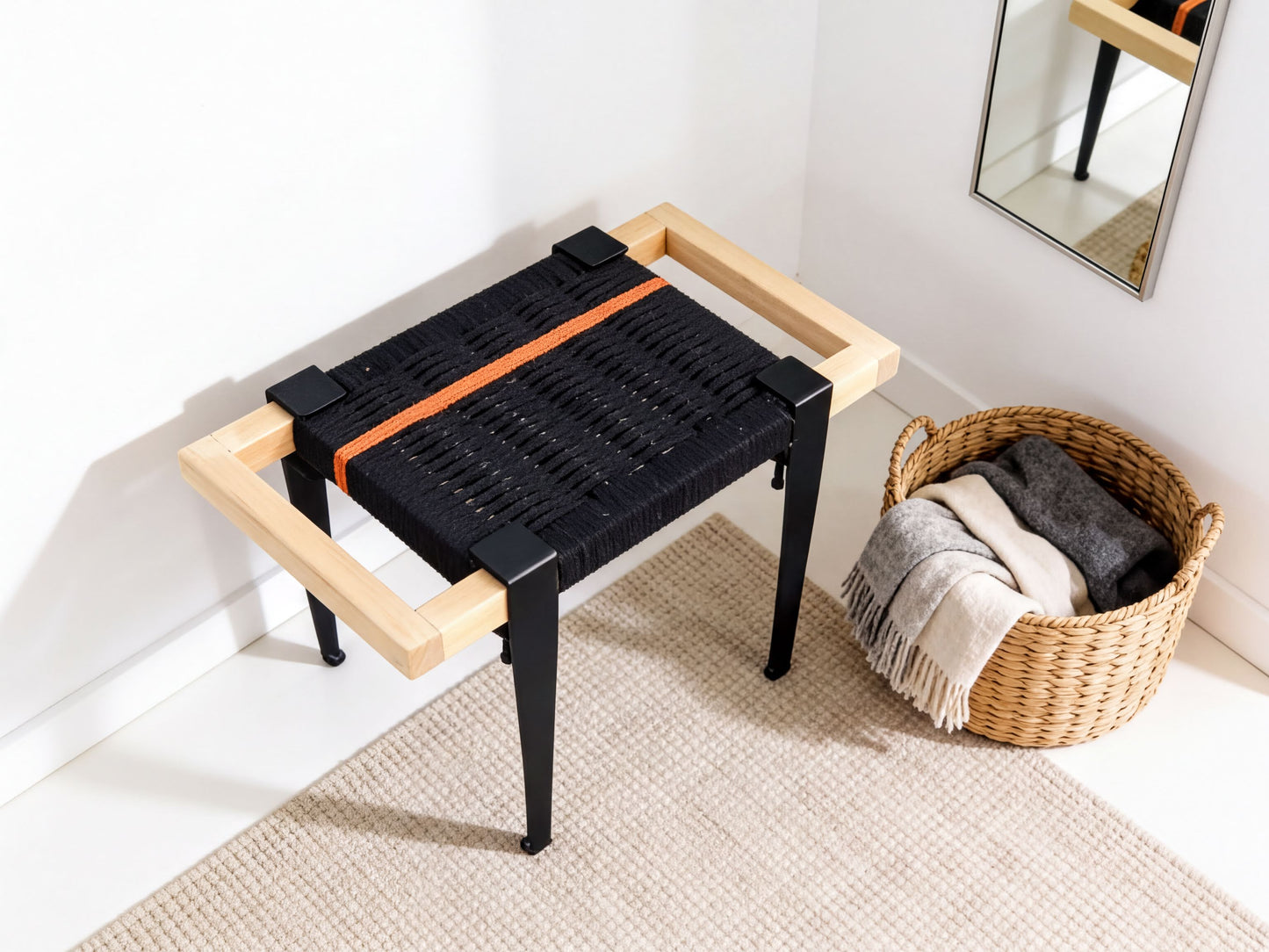 a black woven bench with a wooden frame, placed on a beige rug in a corner of a room. Next to the bench is a woven basket filled with various items, including a gray blanket and a white towel.