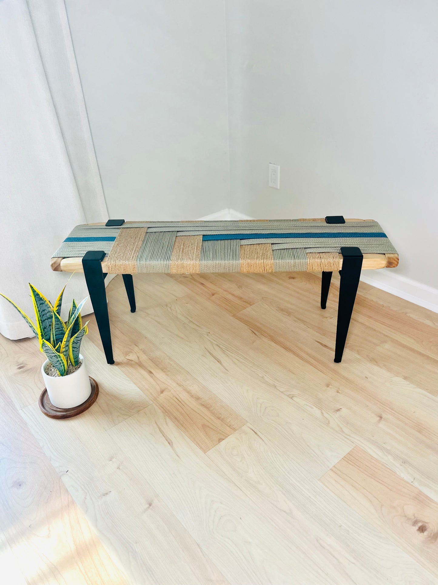 a wooden bench with a woven seat and legs, placed on a wooden floor next to a potted plant.