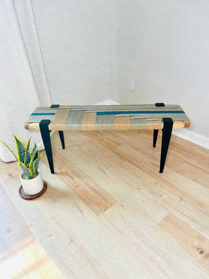 a wooden bench with a woven seat and legs, placed on a wooden floor next to a potted plant.