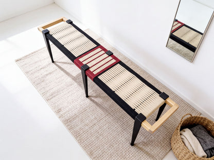 A modern bench with a woven seat and black metal legs, placed on a beige rug in a minimalist room with a mirror and a basket of clothes.