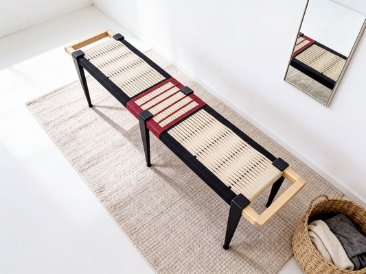 A modern bench with a woven seat and black metal legs, placed on a beige rug in a minimalist room with a mirror and a basket of clothes.