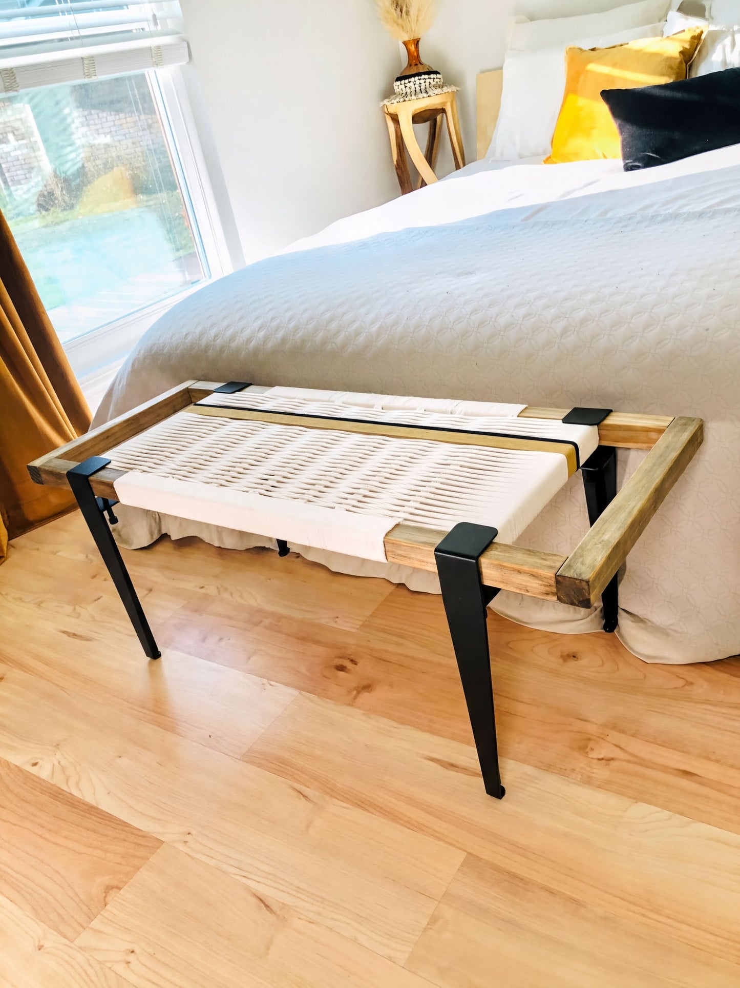 a bedroom with a bed, a bench, and a window. The bench has a white seat and black metal legs, and it is placed in front of the bed. The bed has a gray comforter and yellow pillows, and the room has wooden floors and a window that provides a view of the outside.