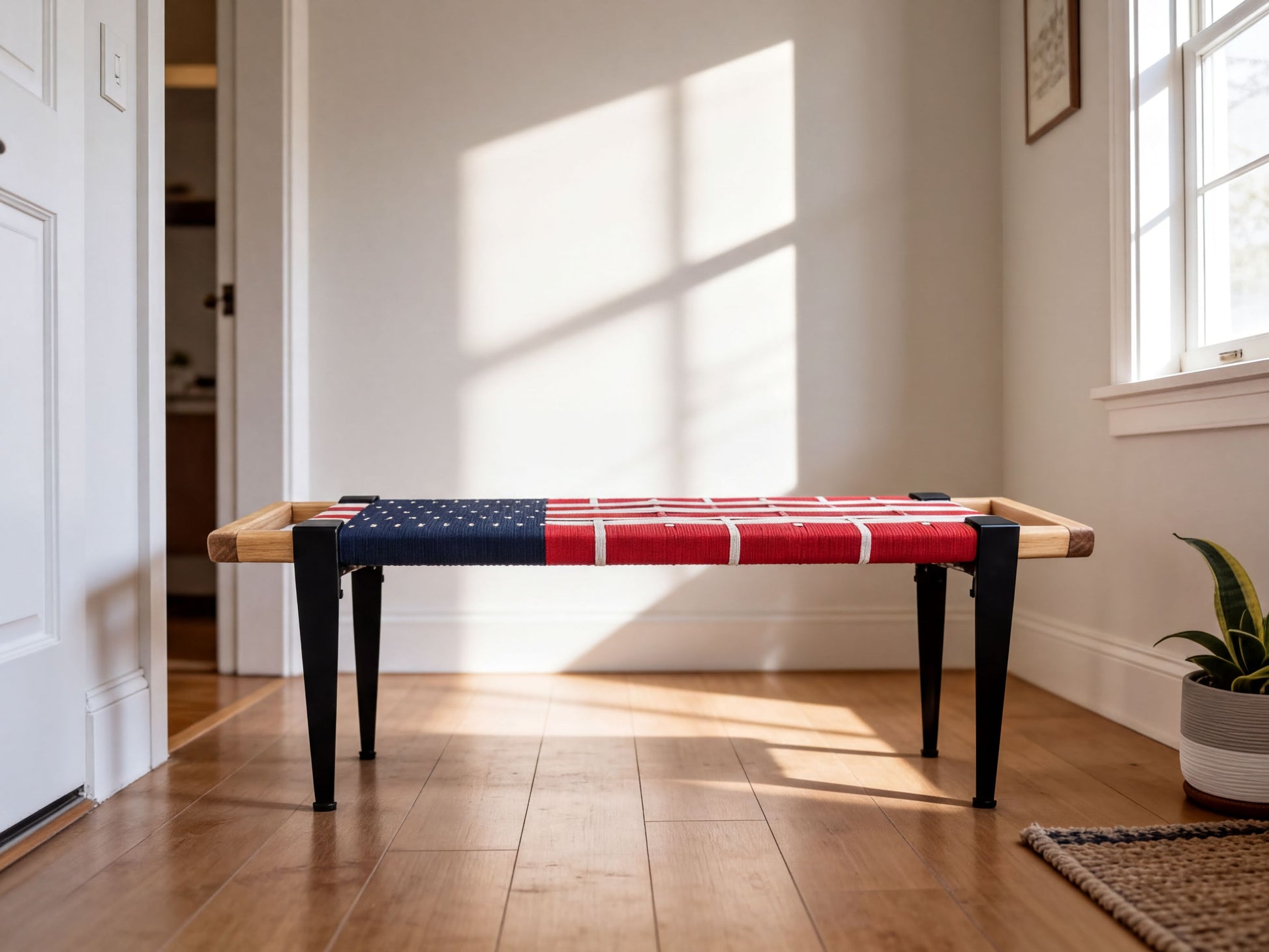 A wooden bench with a red and blue American flag-like design on the seat, placed on a hardwood floor in a room with a white wall and a window.