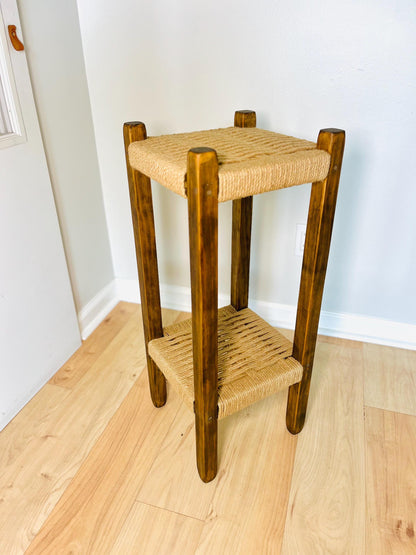A rustic wooden side table with a woven seat and legs, placed on a wooden floor against a white wall.