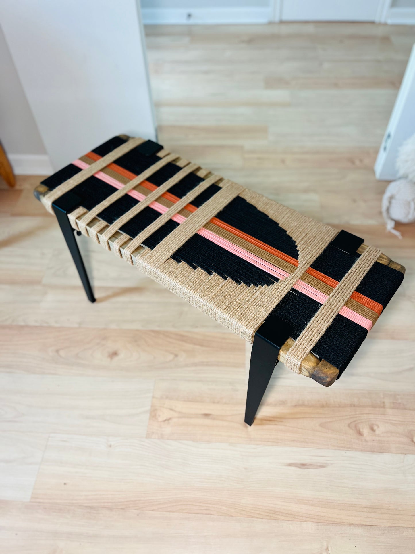 A woven bench with a black and orange design on the seat and legs, placed on a wooden floor.