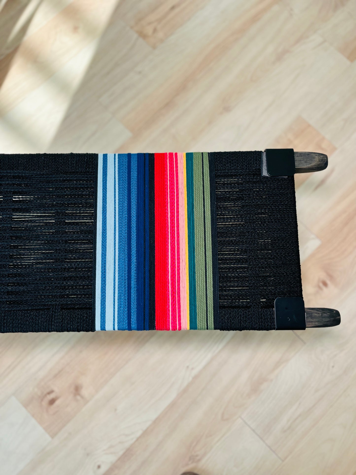 A colorful woven bench with a black seat and wooden legs, featuring a vibrant stripe pattern in red, blue, green, and yellow.