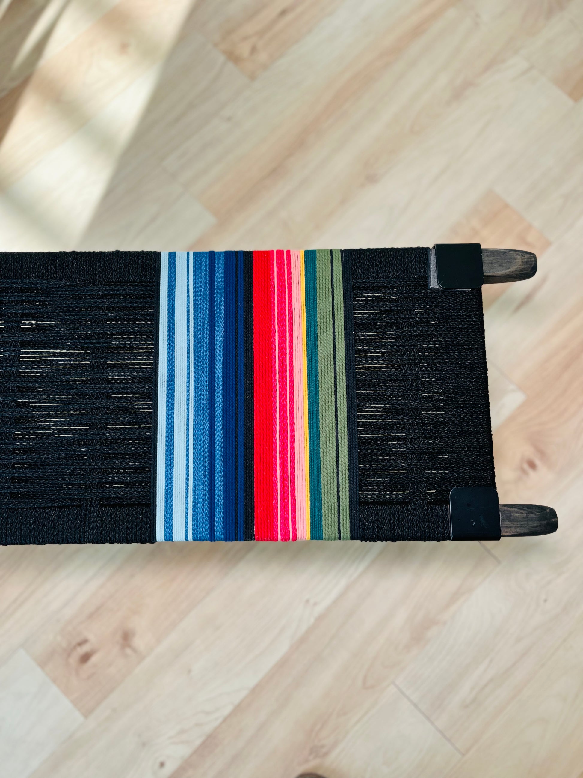 A colorful woven bench with a black seat and wooden legs, featuring a vibrant stripe pattern in red, blue, green, and yellow.