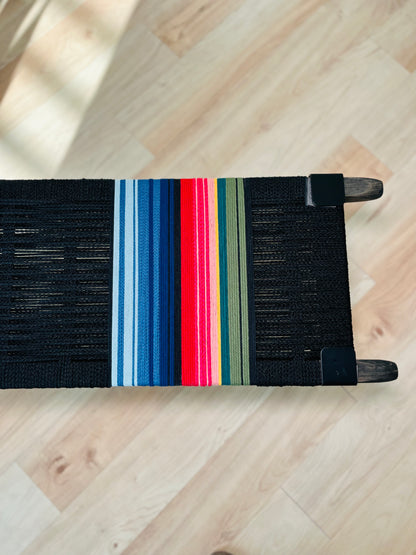 A colorful woven bench with a black seat and wooden legs, featuring a vibrant stripe pattern in red, blue, green, and yellow.