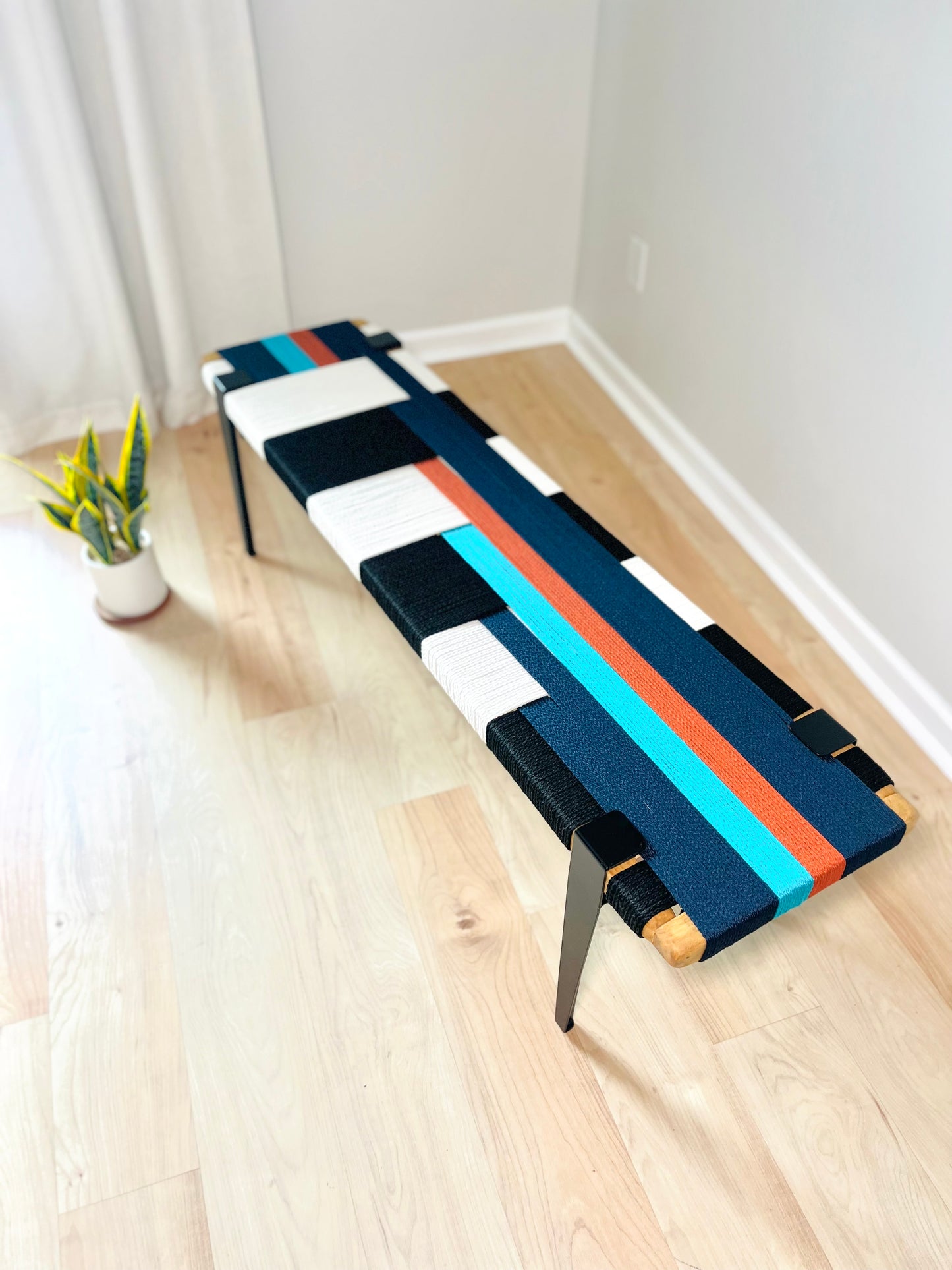a colorful bench with a black and white striped seat, placed on a wooden floor in a room with a white curtain.