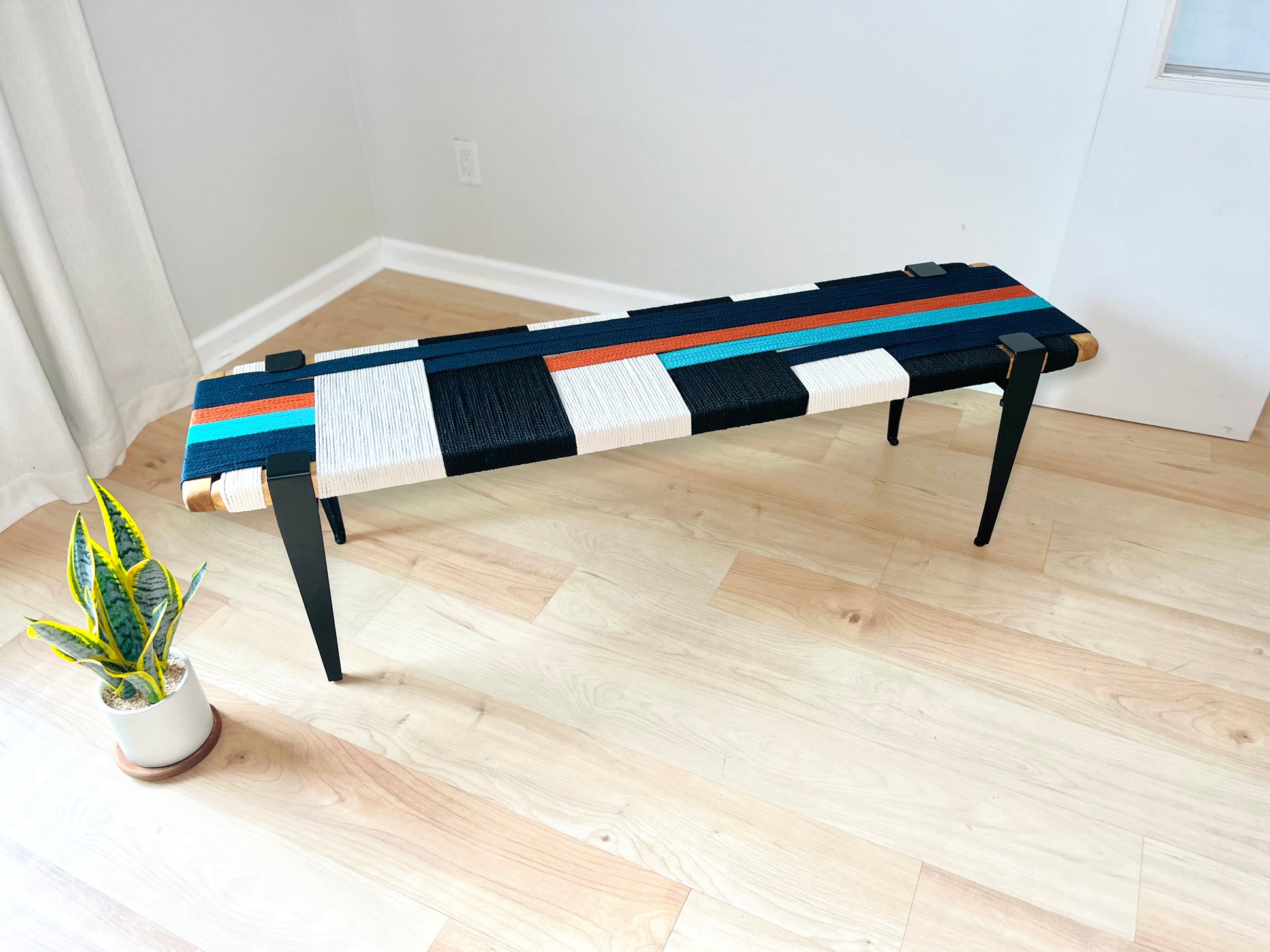 a modern, colorful bench with a black and white striped design, placed on a wooden floor in a room with a white wall and a window.
