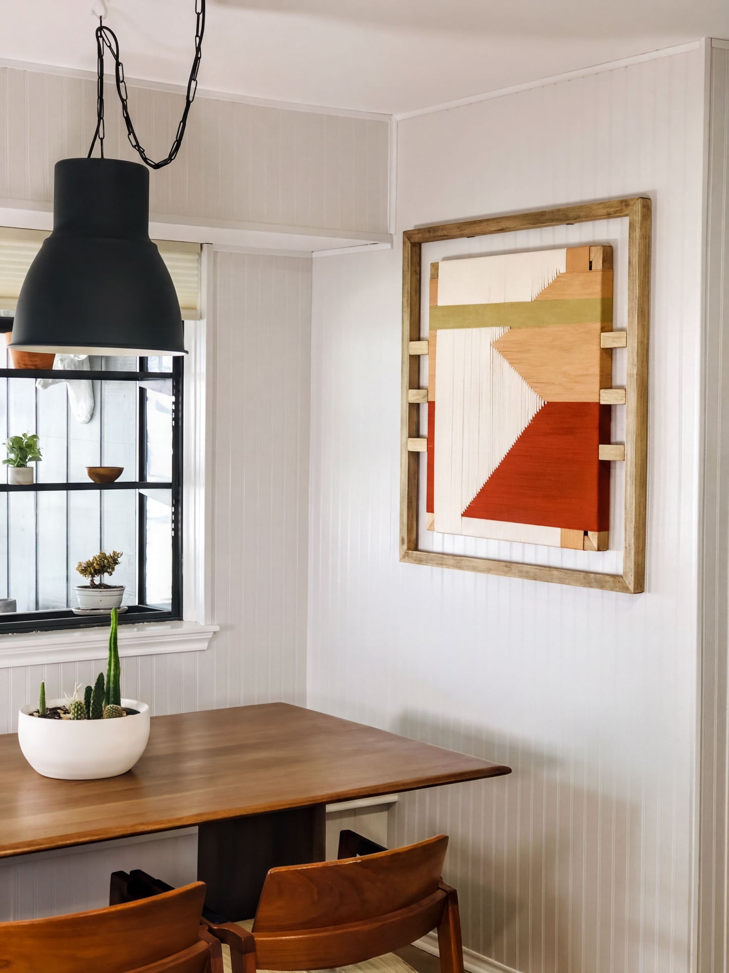 a dining area with a wooden table and chairs, a large abstract painting on the wall, and a potted plant on the windowsill.
