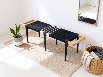 a modern black bench with wooden handles, placed on a beige rug in a room with a mirror and a basket of clothes nearby.