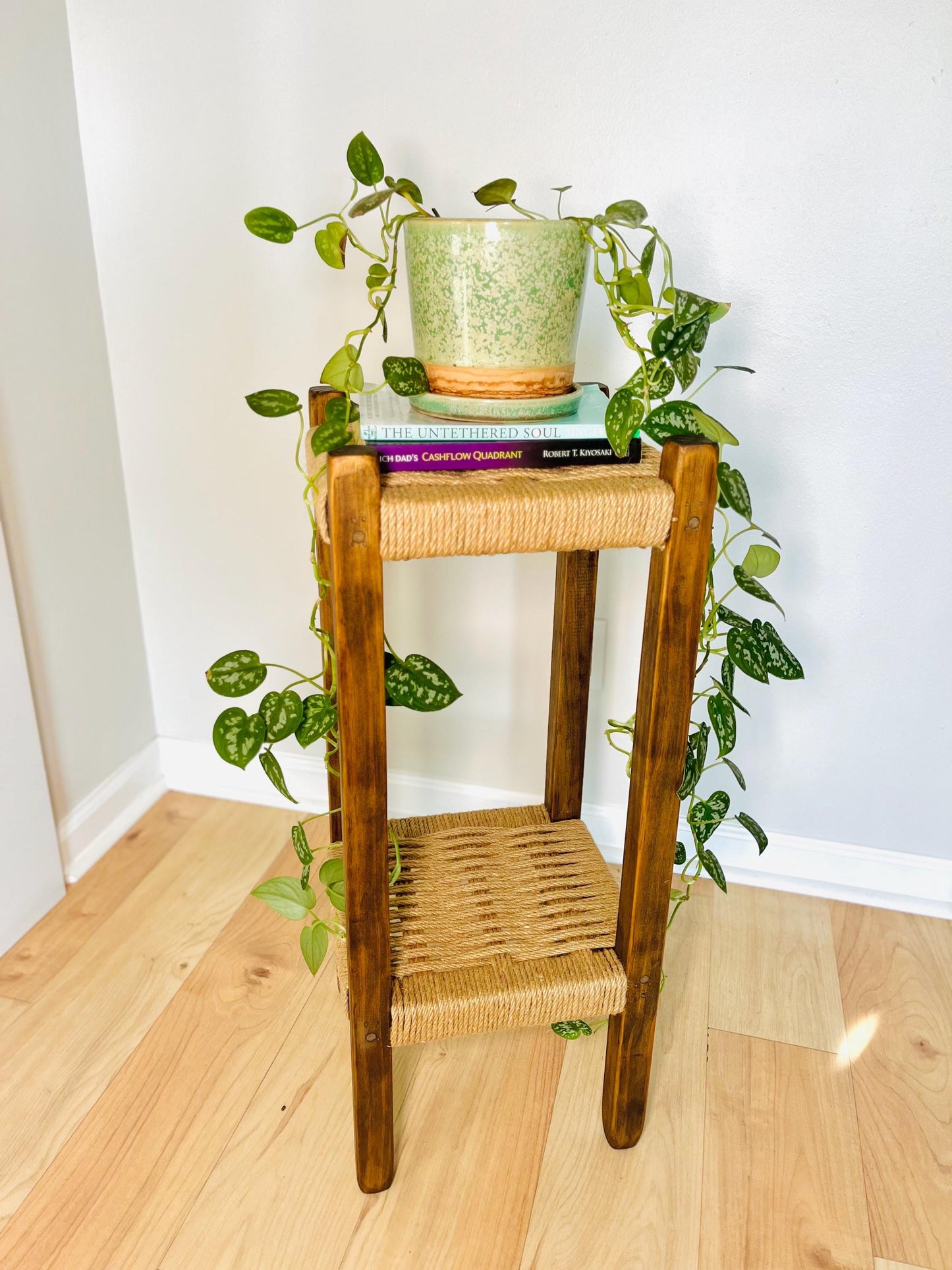 A potted plant with green leaves is placed on a wooden side table, which has a woven seat and is situated on a wooden floor.
