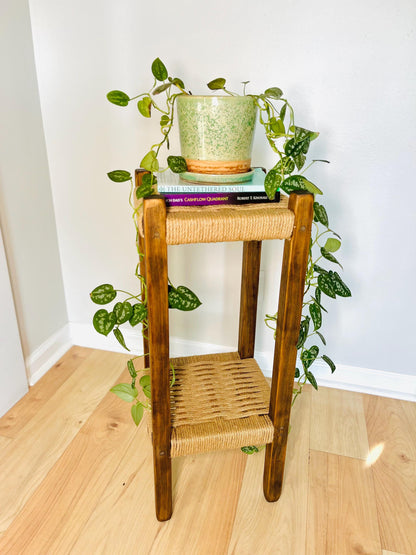A potted plant with green leaves is placed on a wooden side table, which has a woven seat and is situated on a wooden floor.