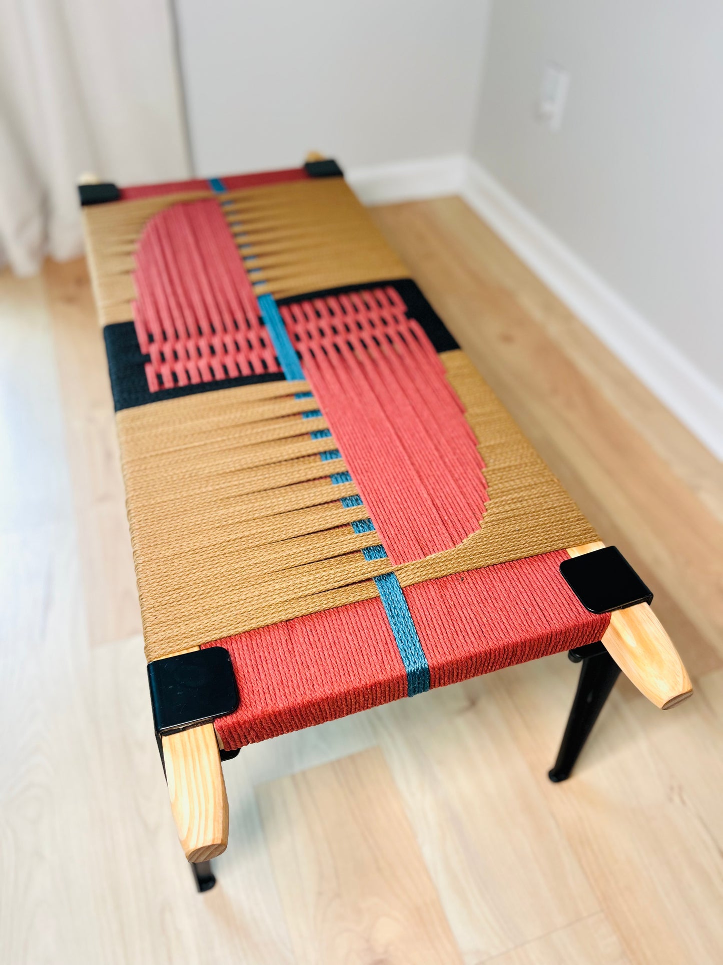a colorful woven bench with a geometric pattern in red, yellow, and black. The bench is placed on a wooden floor against a white wall.