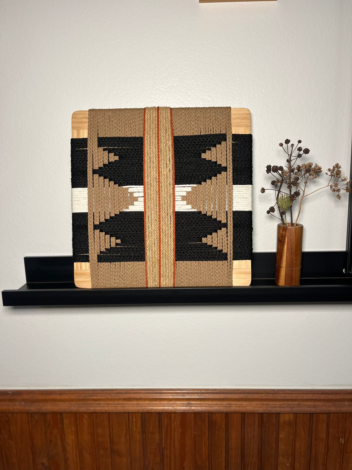 a wooden shelf with a woven piece of art, a vase with dried flowers, and a wooden panel.
