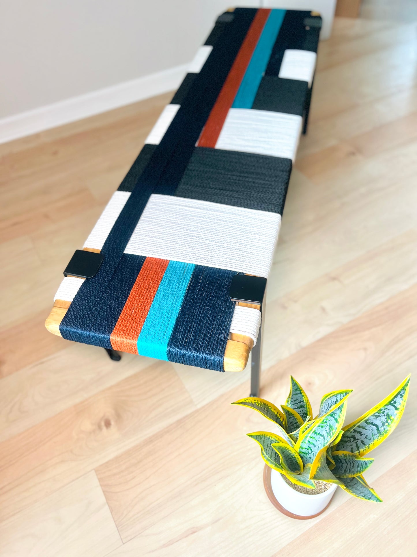 a colorful bench with a black, white, and orange striped seat, placed on a wooden floor next to a potted plant.