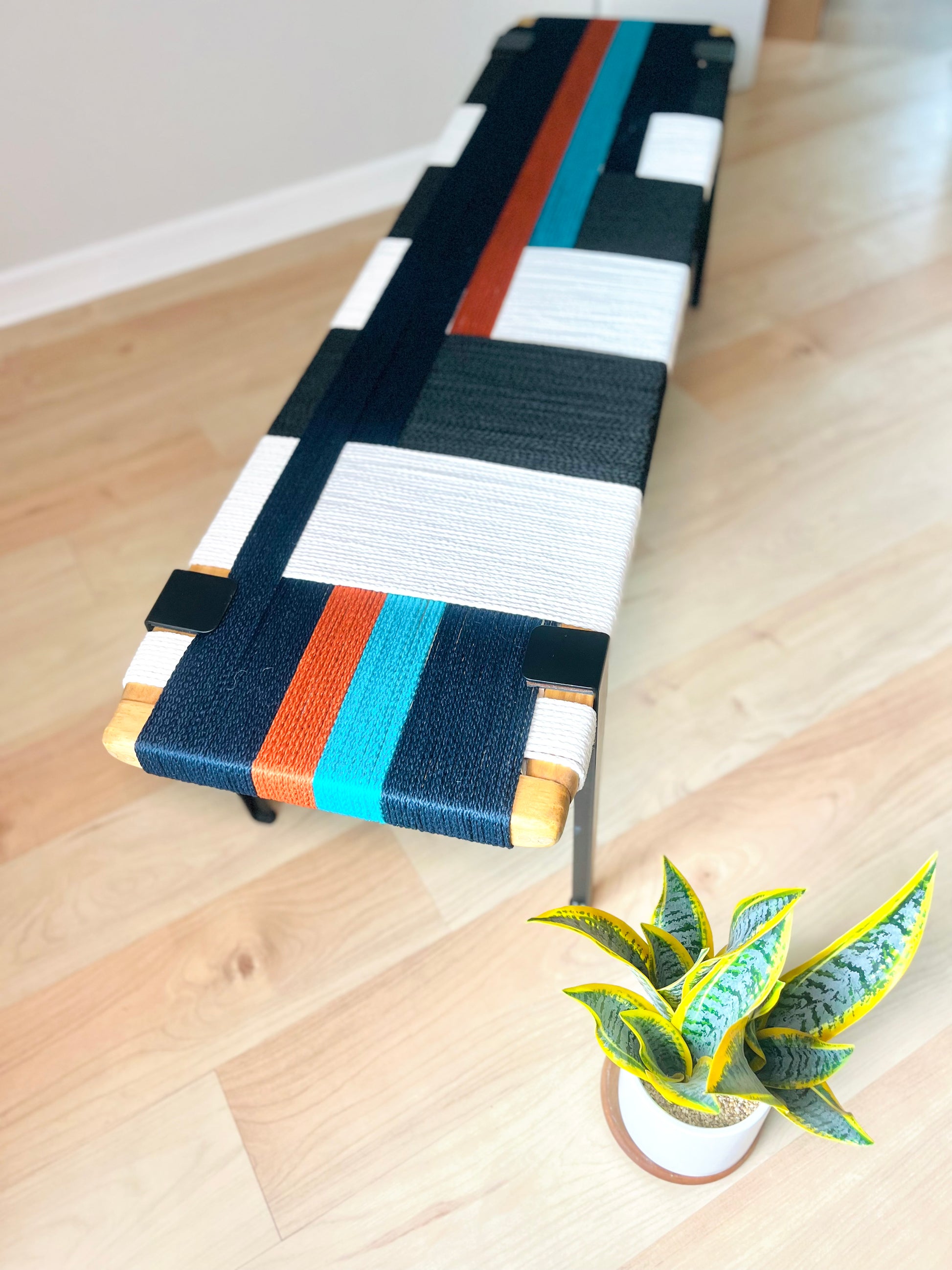 a colorful bench with a black, white, and orange striped seat, placed on a wooden floor next to a potted plant.