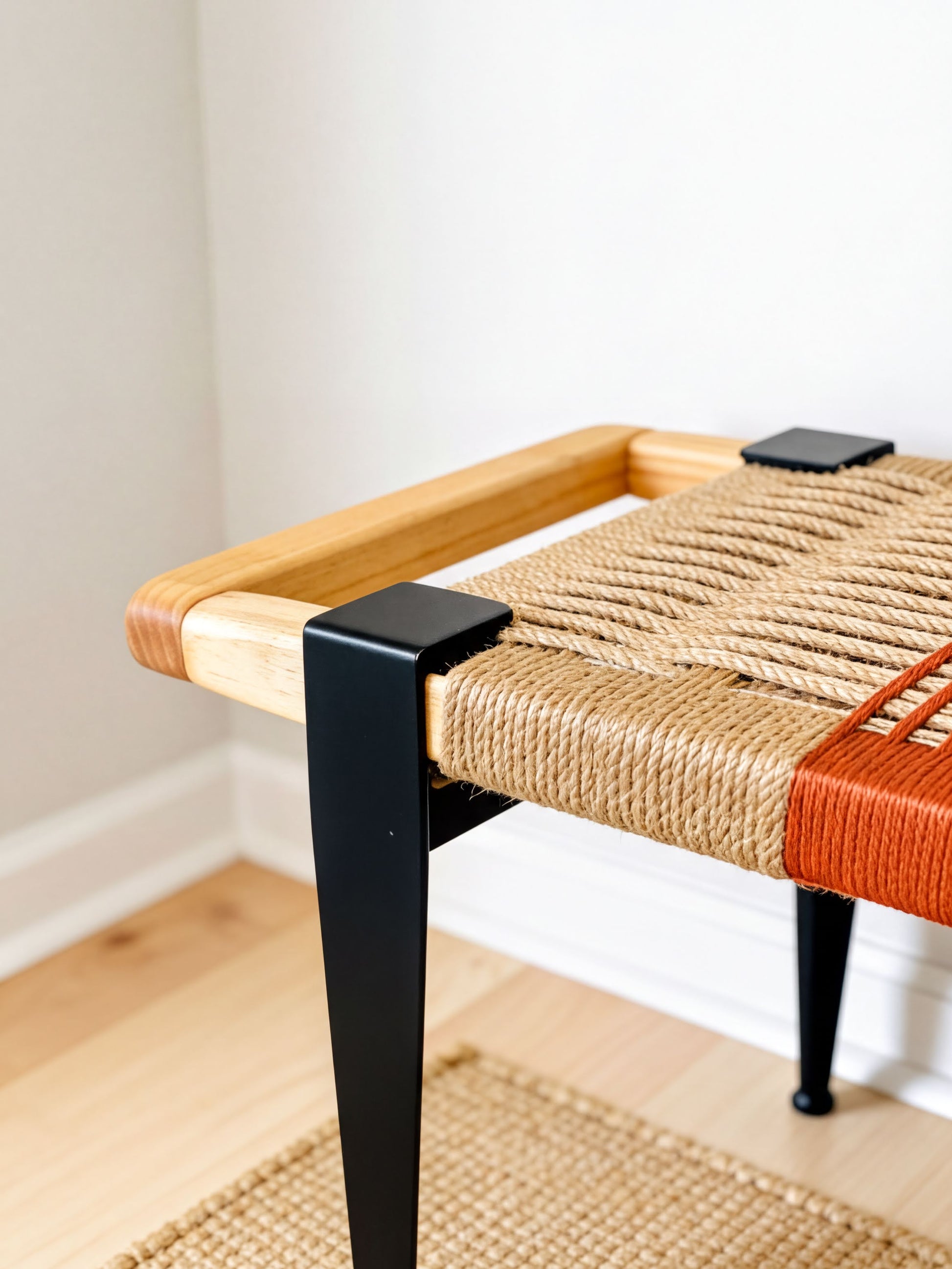 a modern bench with a woven seat and black metal legs, placed on a wooden floor against a white wall.