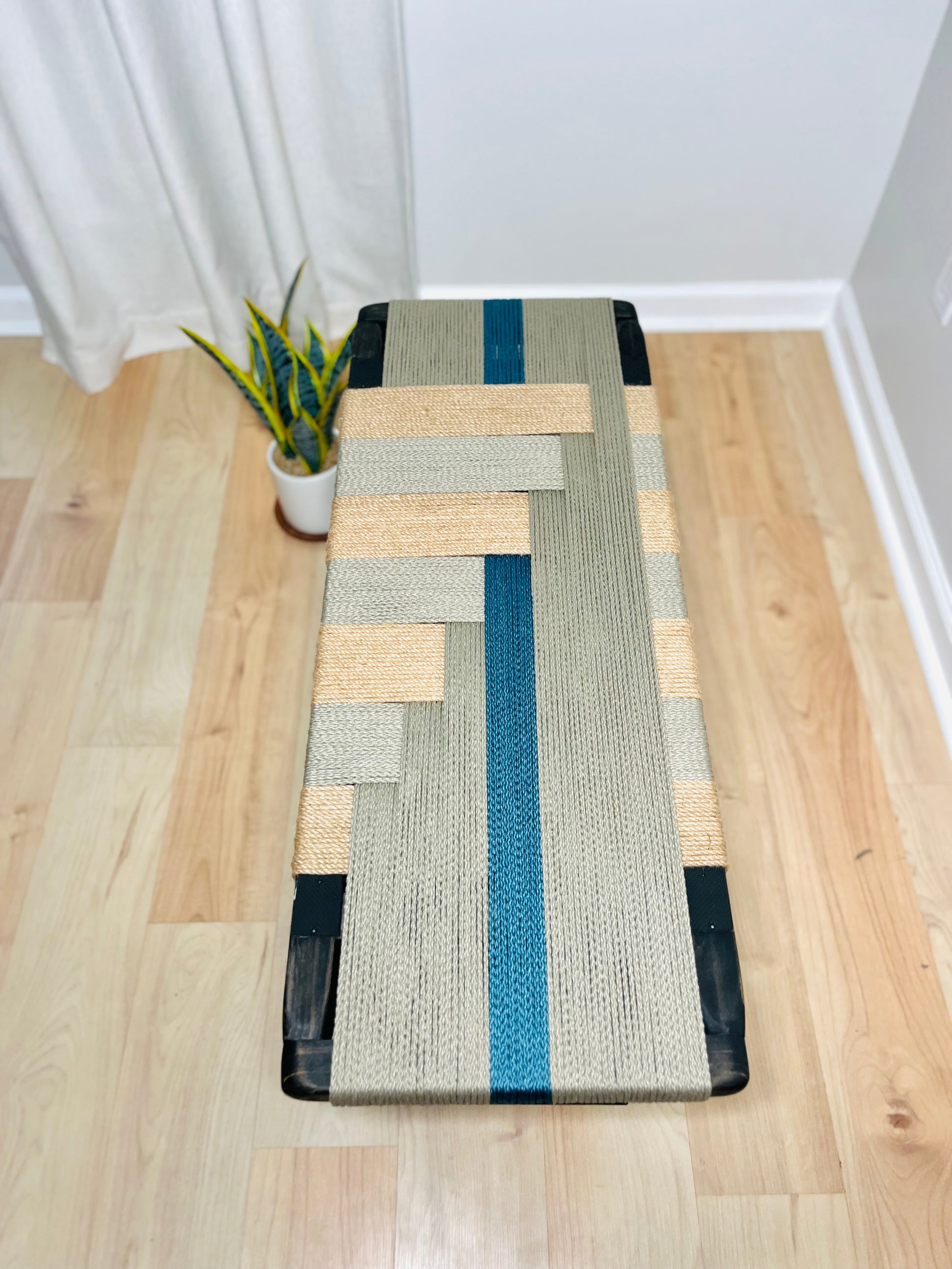 a wooden floor with a woven bench made of a combination of gray and blue fabric, placed next to a potted plant.