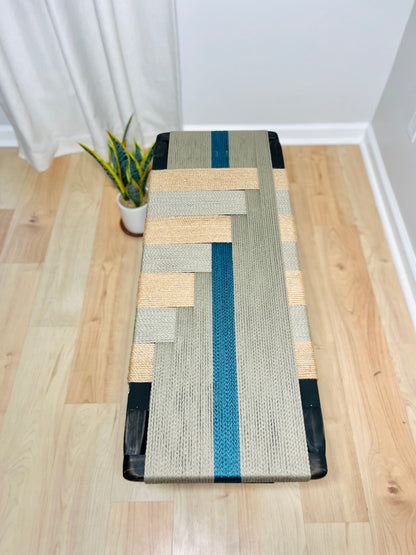 a wooden floor with a woven bench made of a combination of gray and blue fabric, placed next to a potted plant.