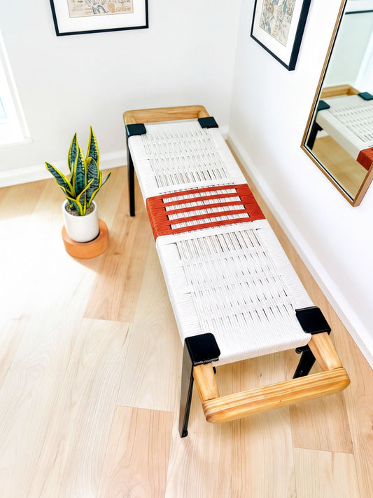a modern, minimalist living space with a wooden bench and a potted plant. The bench has a white woven seat and black metal frame, and it is placed on a wooden floor. The potted plant is located next to the bench, adding a touch of greenery to the room. The overall design and color scheme of the space create a clean and contemporary aesthetic.
