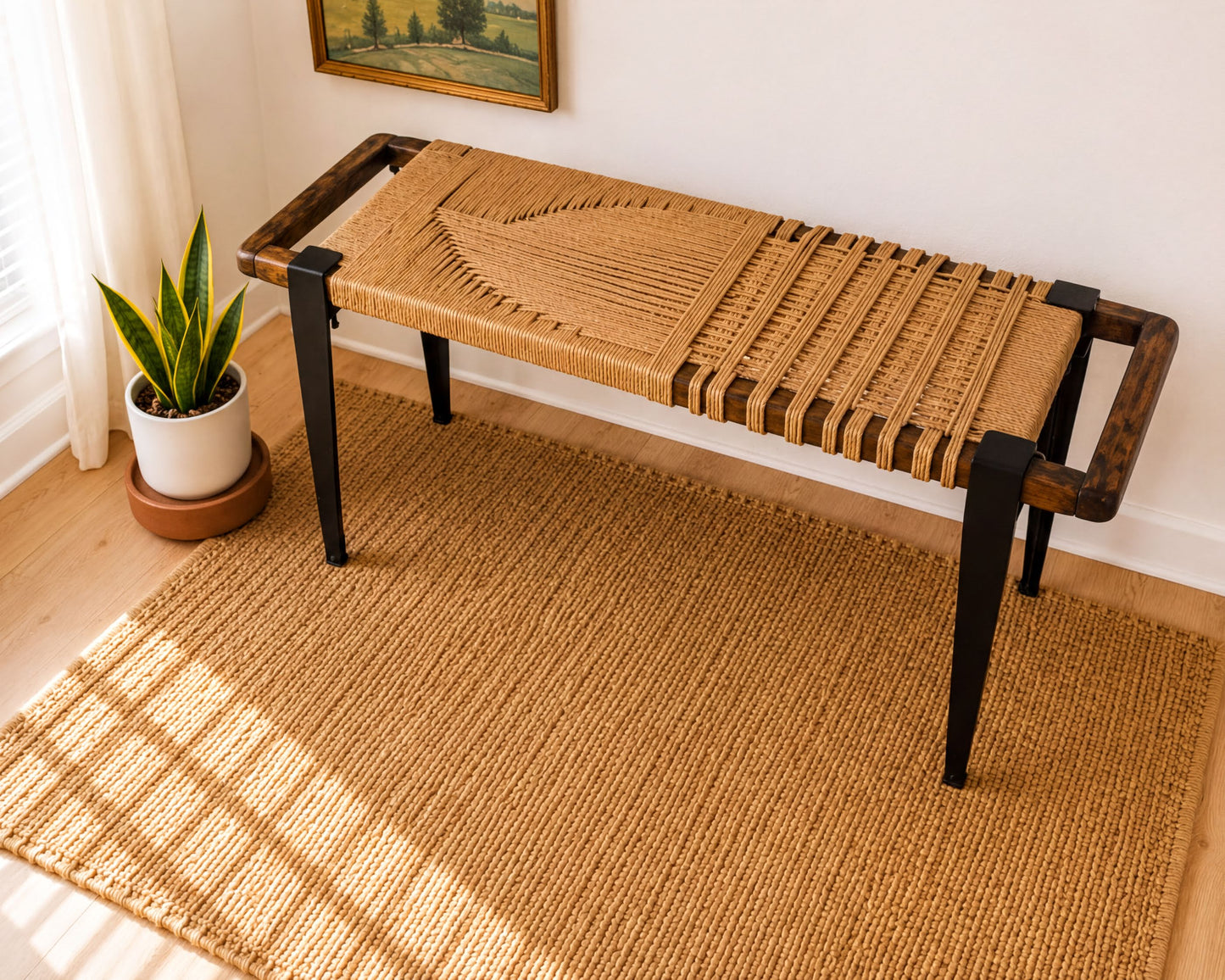 a woven bench with a woven seat and wooden legs, placed on a woven rug in a room with a painting on the wall and a potted plant nearby.