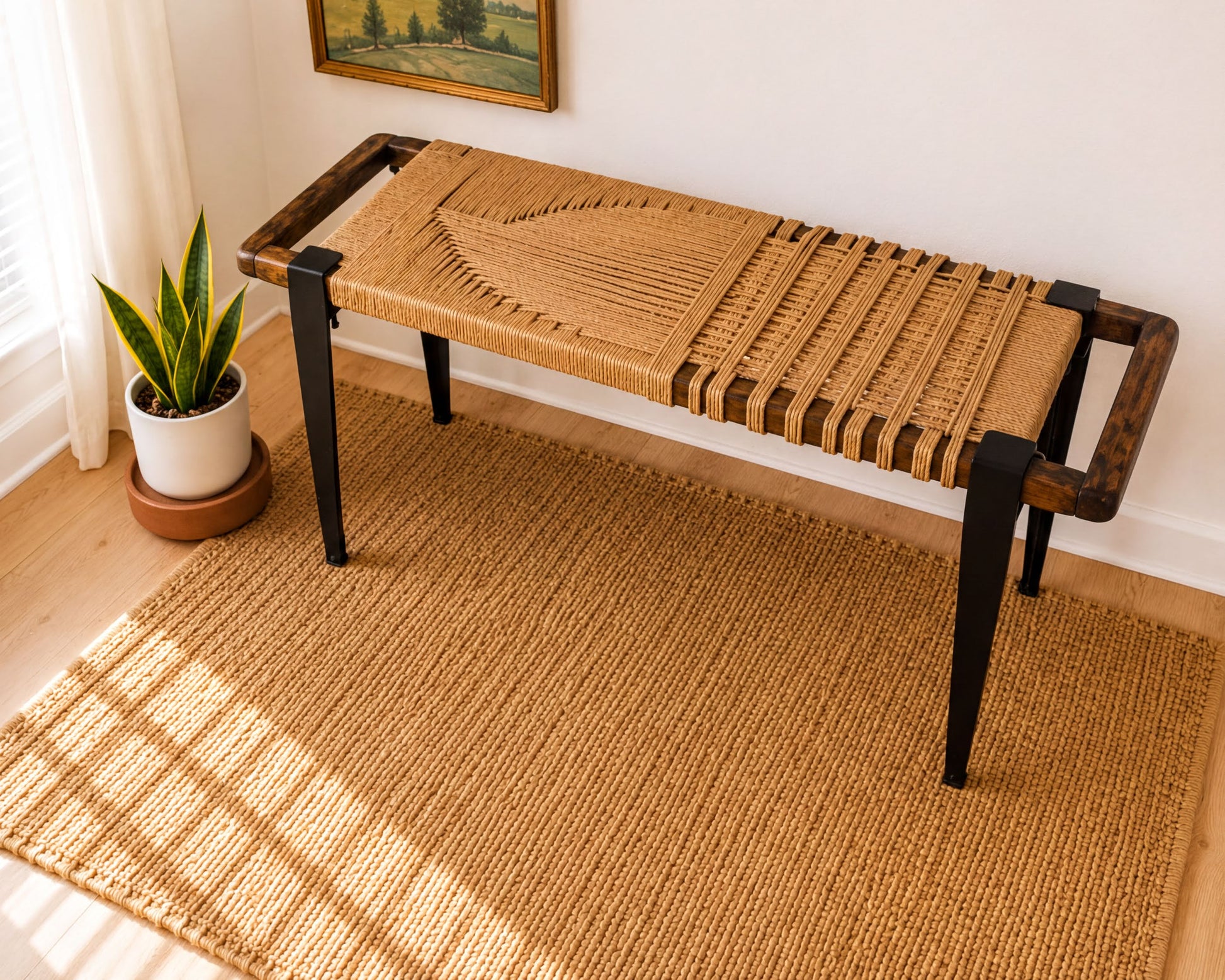 a woven bench with a woven seat and wooden legs, placed on a woven rug in a room with a painting on the wall and a potted plant nearby.