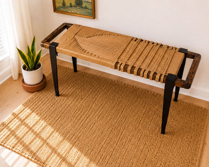 a woven bench with a woven seat and wooden legs, placed on a woven rug in a room with a painting on the wall and a potted plant nearby.