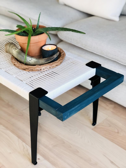 a modern, minimalist coffee table with a white woven base and a blue frame. On the table, there is a potted plant, a candle, and a small sculpture. The table is placed on a wooden floor, and there is a couch visible in the background.