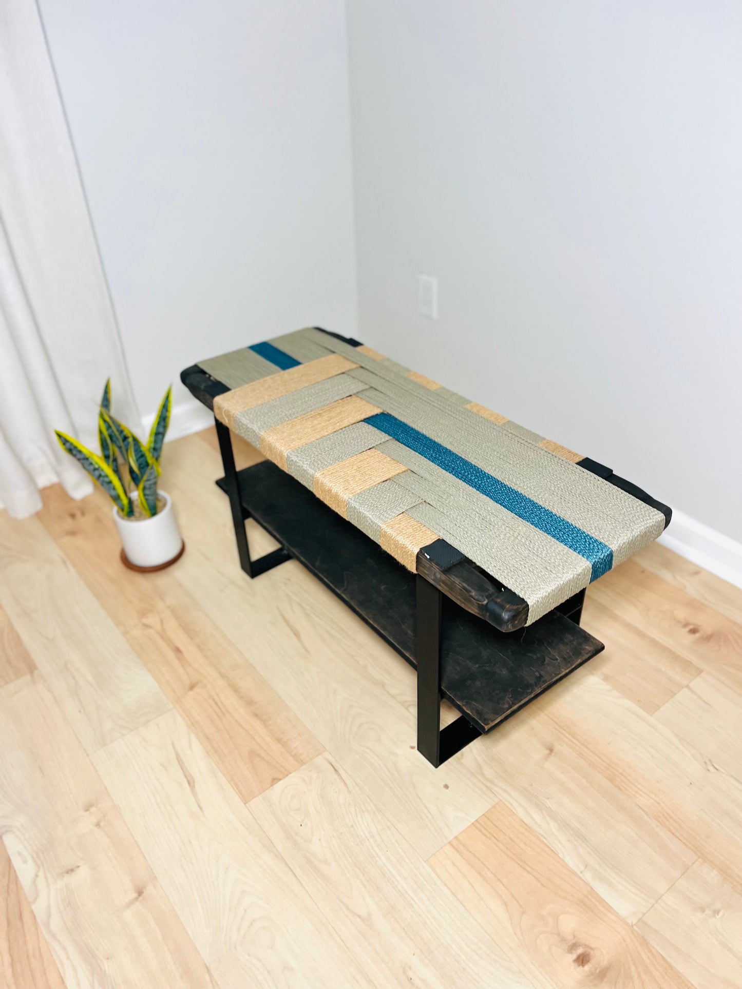 a modern bench made of woven materials, placed on a wooden floor in a room with a white curtain.