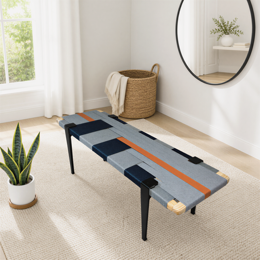 a modern living room with a unique bench-style table with a geometric pattern in shades of blue, orange, and gray. The table is placed on a beige rug, and there is a round mirror hanging on the wall above it. A woven basket is also present in the room, and a potted plant is visible on the left side of the image.