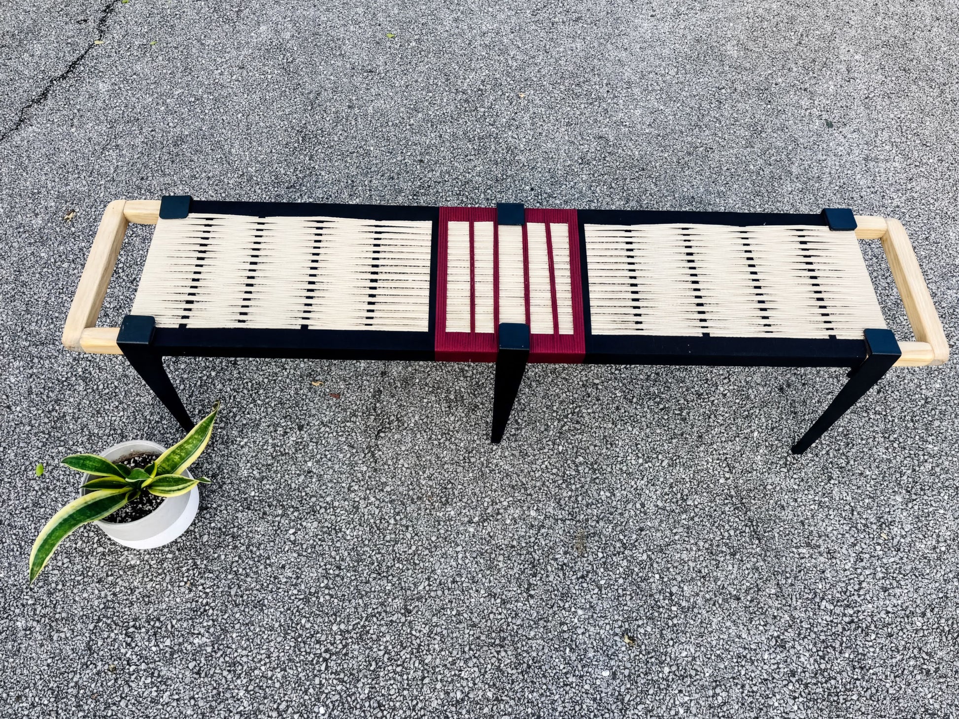 a bench with a black and red frame, a white seat, and two wooden handles on either side. The bench is placed on a gray surface, and there is a potted plant nearby.