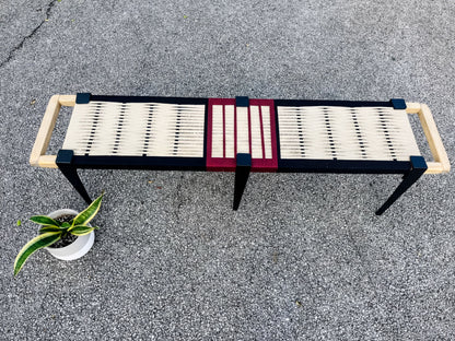 a bench with a black and red frame, a white seat, and two wooden handles on either side. The bench is placed on a gray surface, and there is a potted plant nearby.