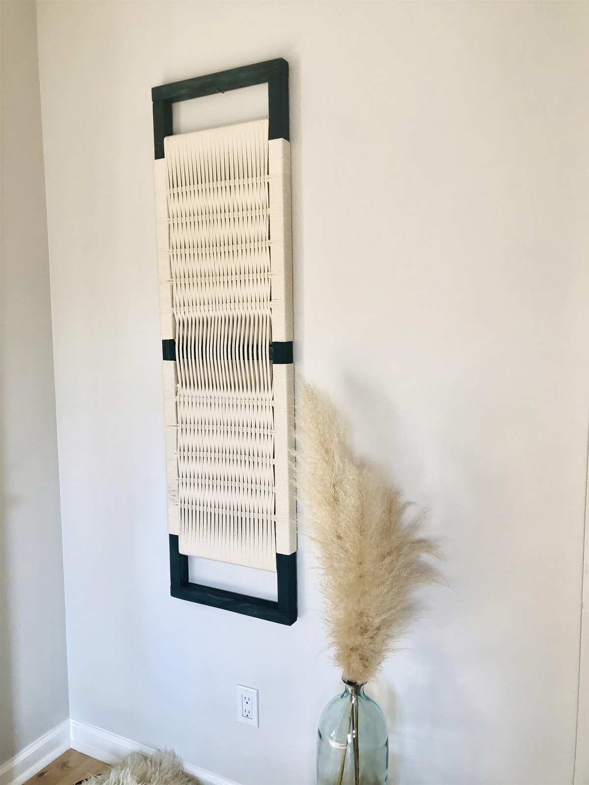 a white woven wall hanging with a black frame, a glass vase with a potted plant, and a white wall.
