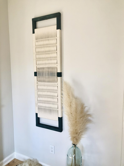 a white woven wall hanging with a black frame, a glass vase with a potted plant, and a white wall.