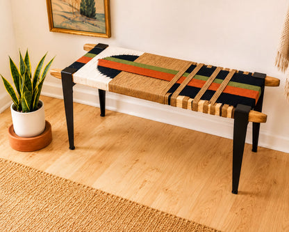 a wooden bench with a colorful woven seat, placed on a wooden floor next to a potted plant.