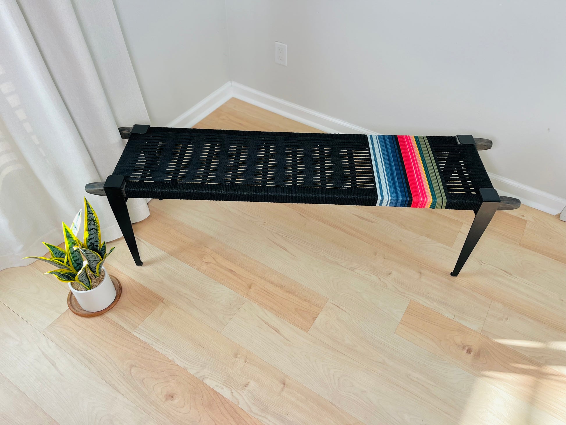 a black bench with a colorful striped design on the seat, placed on a wooden floor next to a potted plant.